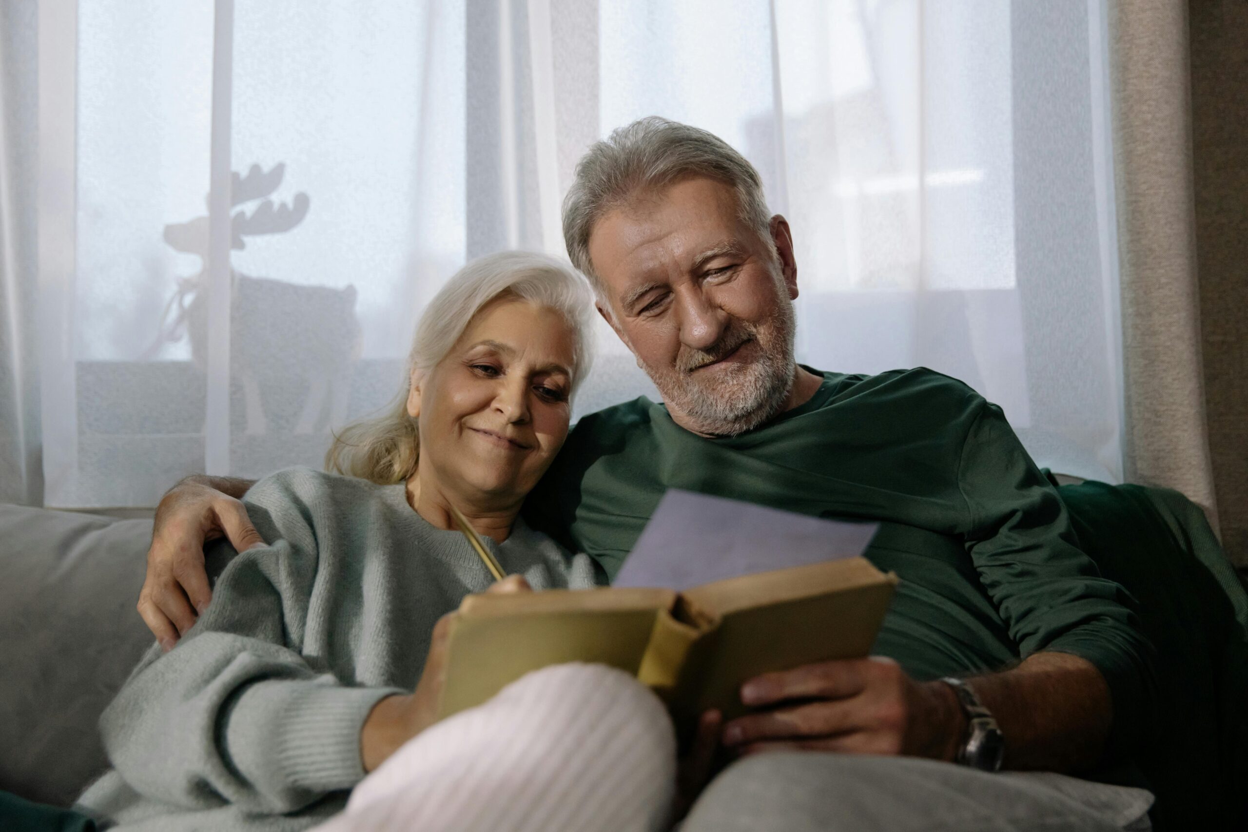 Elderly couple sitting close together reading a book at home.